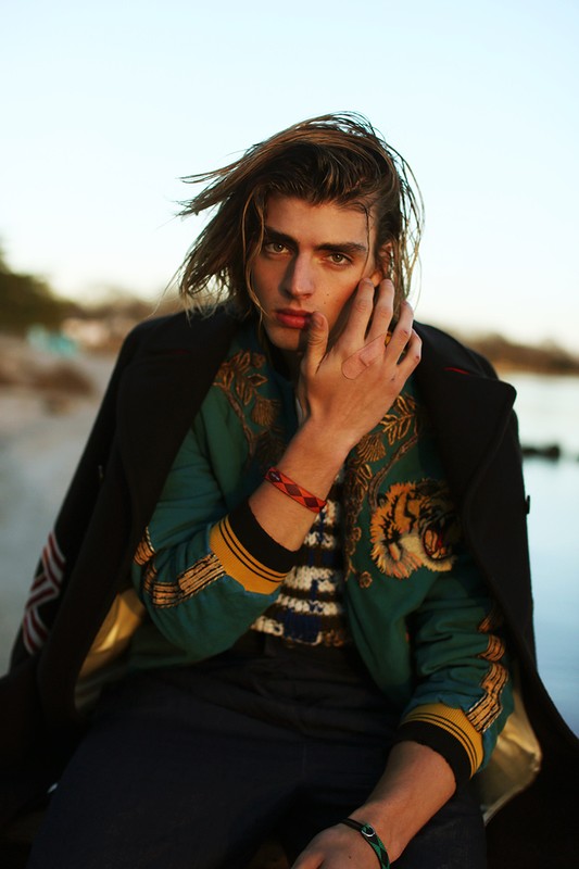 Portrait of a young adult male model with long blond hair, wearing an embroidered jacket and blue jeans, posing outdoors by the water.