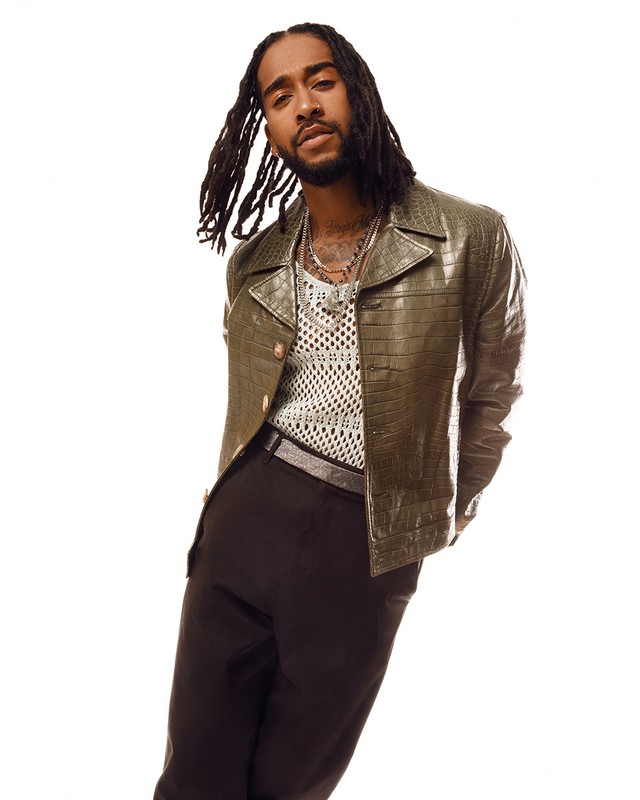 Portrait of a stylish adult male with dreadlocks wearing an olive green reptile-pattern jacket and trousers against a white background.