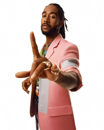 Portrait of Omarion wearing a pink blazer with grey panels, holding his hand toward the camera against a white background