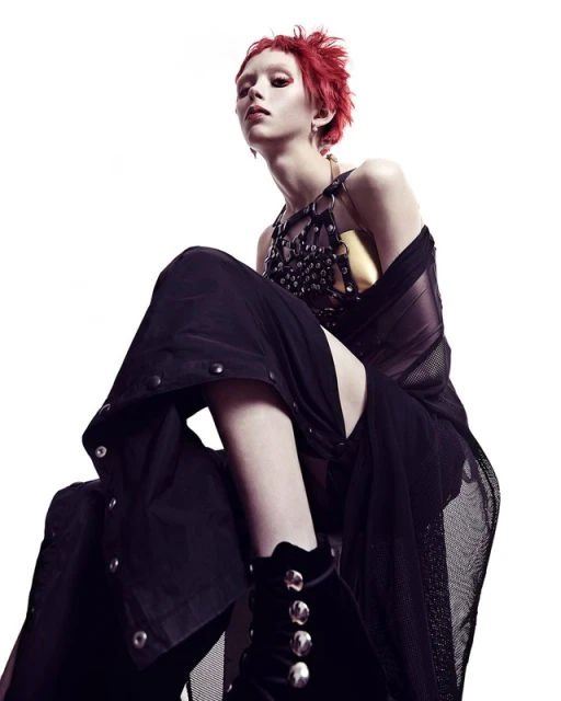 Red-haired person in a dark, avant-garde outfit featuring a studded harness, mesh top, and velvet gloves posing against a white backdrop.