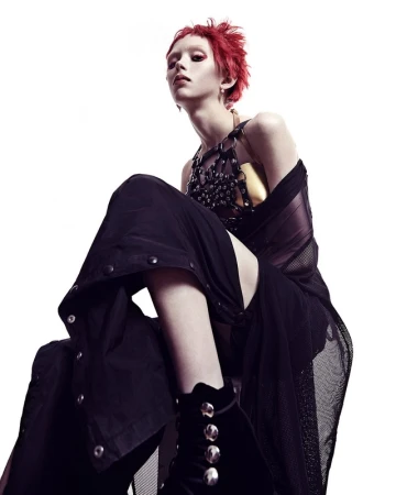 Red-haired person in a dark, avant-garde outfit featuring a studded harness, mesh top, and velvet gloves posing against a white backdrop.