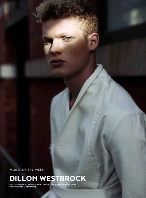 Portrait of a light-skinned adult male model with curly hair wearing a white robe against a red brick urban background in natural light.