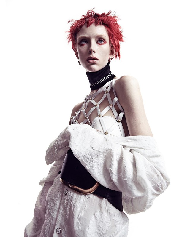 A model with fiery red hair and intense eye makeup poses in a white, textured shirt and structured top against a bright white backdrop.