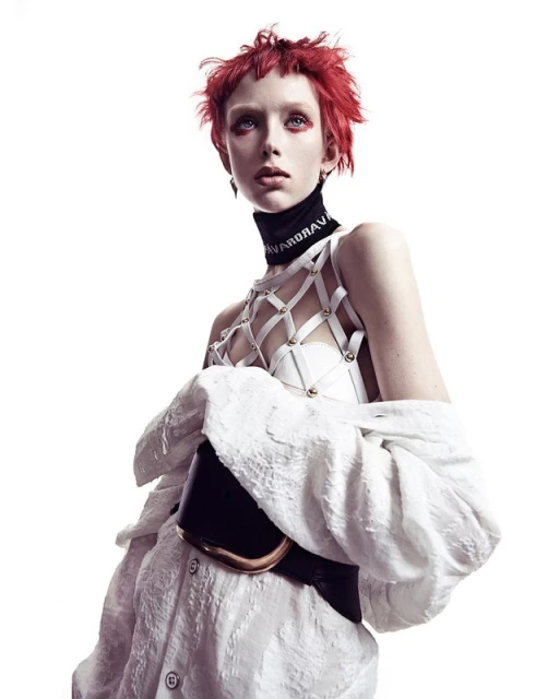 A model with fiery red hair and intense eye makeup poses in a white, textured shirt and structured top against a bright white backdrop.
