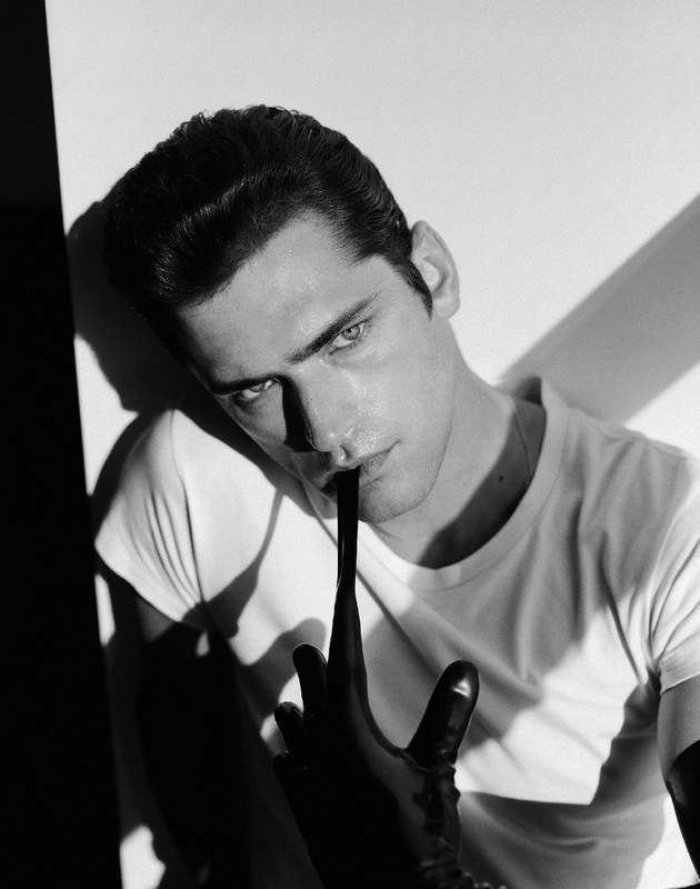 Black and white portrait of a fair-skinned adult male in a white t-shirt, holding a black latex glove to his mouth in a studio.