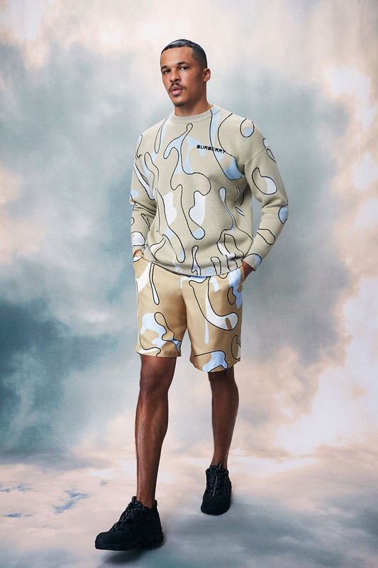 A male model poses in a studio, wearing a matching beige and light blue camo pattern sweater and shorts with black shoes against a cloudy backdrop.