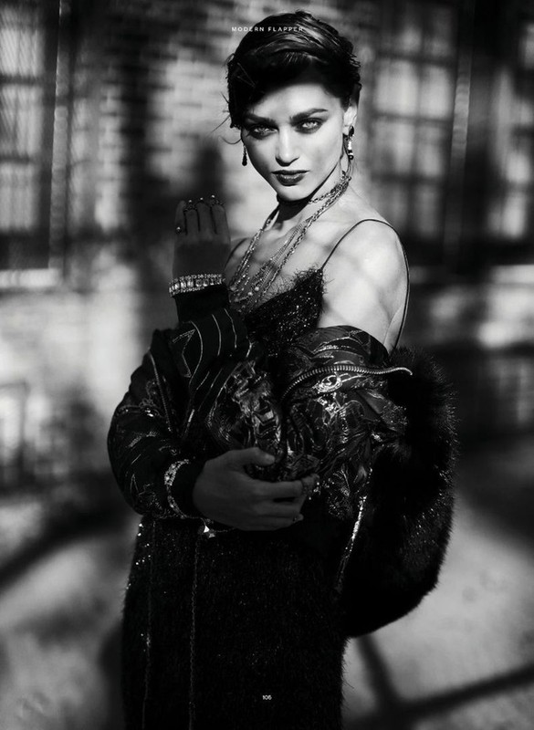 A monochrome fashion portrait of a woman wearing a dark dress, fur coat, and jewelry, posed against a shadowed background.