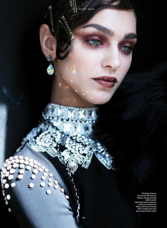 Close-up portrait of a brunette woman with elaborate makeup, wearing a jeweled collar and multiple bobby pins in her curled hair.