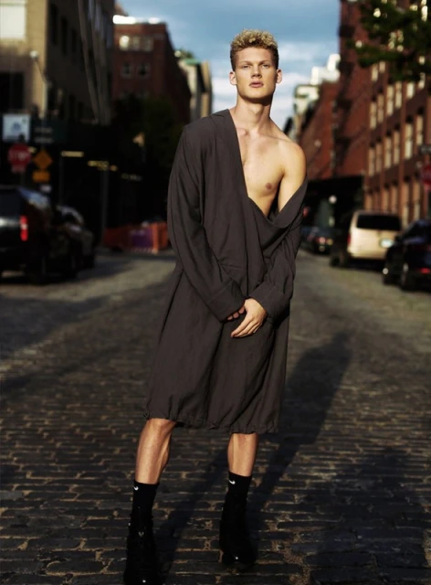 A young adult male stands on a cobblestone city street in a long, open grey shirt with black socks and dress shoes, posing fashionably.