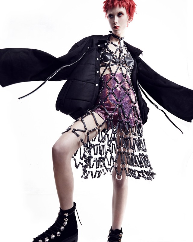 Fashionable young adult woman with red hair poses in edgy outfit in bright studio. She is wearing a black coat and studded boots.