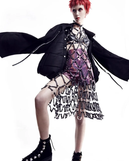 Fashionable young adult woman with red hair poses in edgy outfit in bright studio. She is wearing a black coat and studded boots.