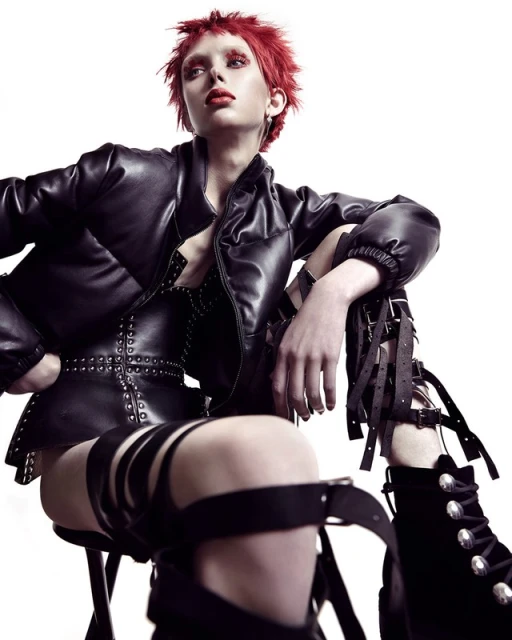 A woman with short, red hair sits on a stool wearing a black leather jacket and a corset-style top with thigh-high boots in a photo studio.