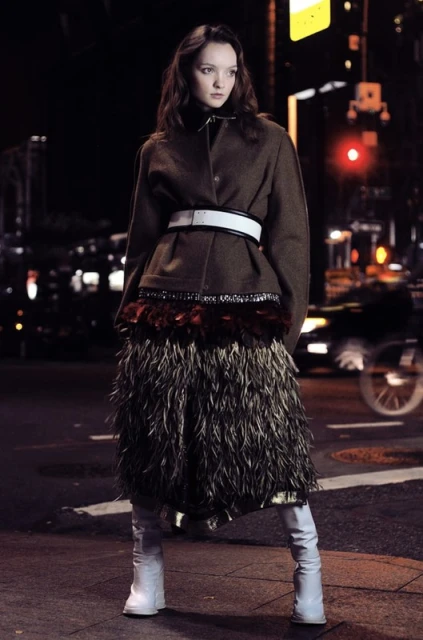 A young woman in a brown jacket and feather skirt stands on a city street at night, illuminated by streetlights and car headlights.
