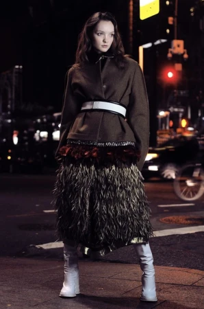 A young woman in a brown jacket and feather skirt stands on a city street at night, illuminated by streetlights and car headlights.