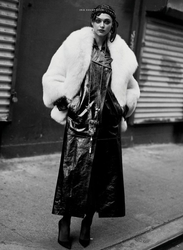 A woman wearing a white faux fur coat and black shiny trench coat stands on a city street in a black and white fashion photograph.