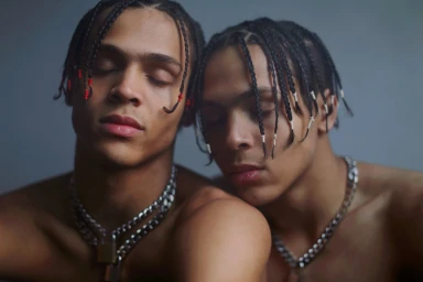 Two young men with braided hair and silver chain necklaces posing closely together with closed eyes.