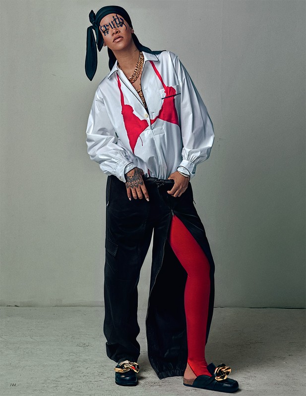 Fashion portrait of a woman with face art wearing a white collared shirt, black pants, a headscarf, and bold jewelry posing indoors.
