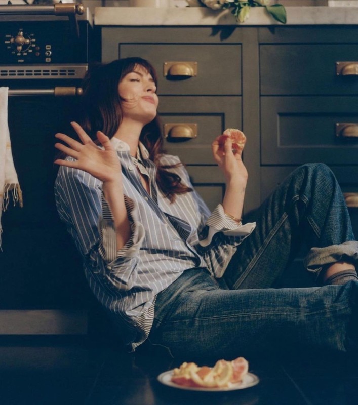Woman in striped shirt and jeans sits on kitchen floor eating citrus fruit, with cabinets and oven visible in the background.