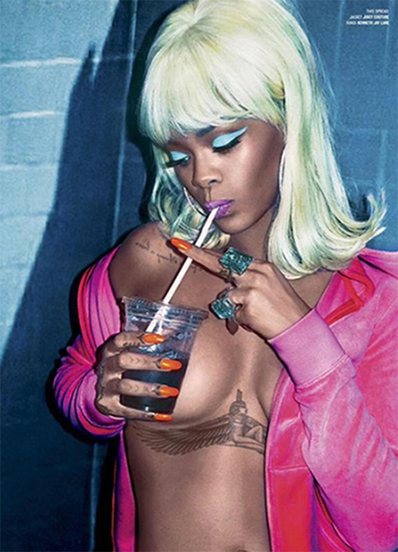 Close-up portrait of a young adult female with blonde hair drinking from a plastic cup, wearing a pink top and rings, against a blue wall.
