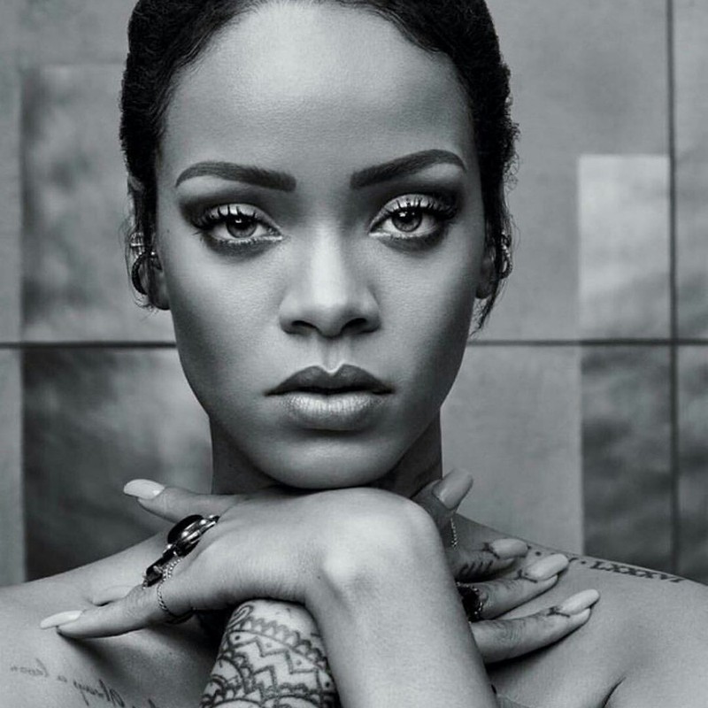 Close-up monochrome portrait of Rihanna with hands crossed under her chin, showcasing makeup, jewelry, and tattoos against a neutral background.