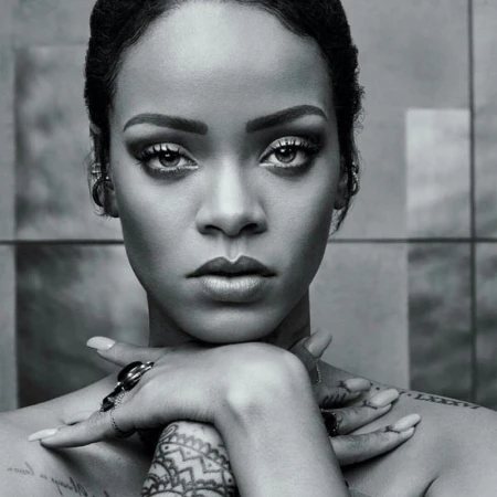 Close-up monochrome portrait of Rihanna with hands crossed under her chin, showcasing makeup, jewelry, and tattoos against a neutral background.