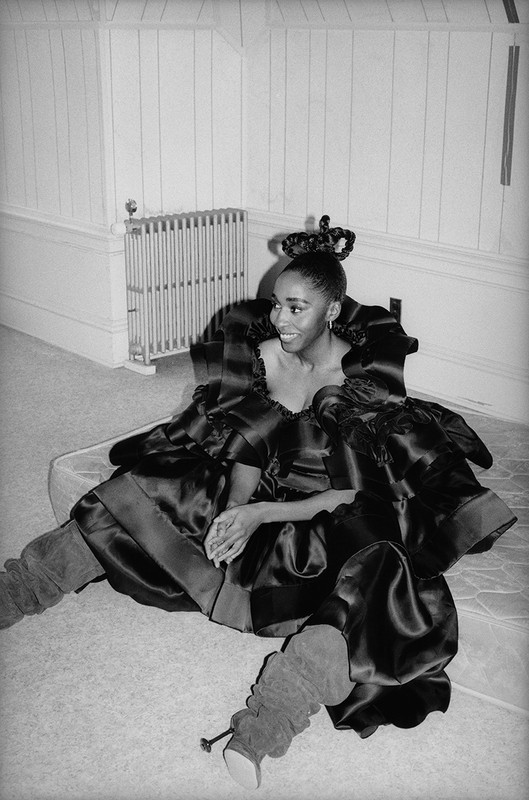 A woman in a large black dress and leg warmers sits on a mattress on the floor, smiling in a monochrome indoor setting.