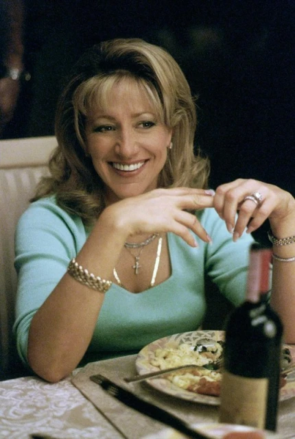 Smiling woman with blonde hair sitting at a table with a plate of food and a bottle of wine, looking off to the side.