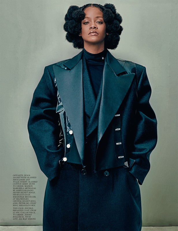 A woman with styled afro hair and a black jacket poses in a studio against a neutral backdrop for a fashion editorial shot.