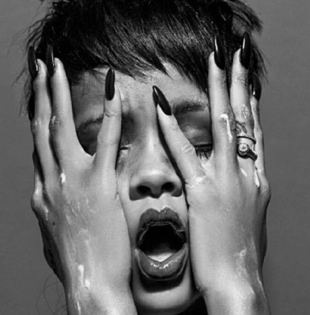Close-up of Rihanna with hands framing her face, showcasing long dark stiletto nails and rings.