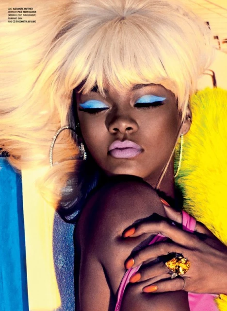 Close-up portrait of a woman with blonde hair, bright blue eye makeup, and lilac lipstick, wearing a pink top and large hoop earrings.