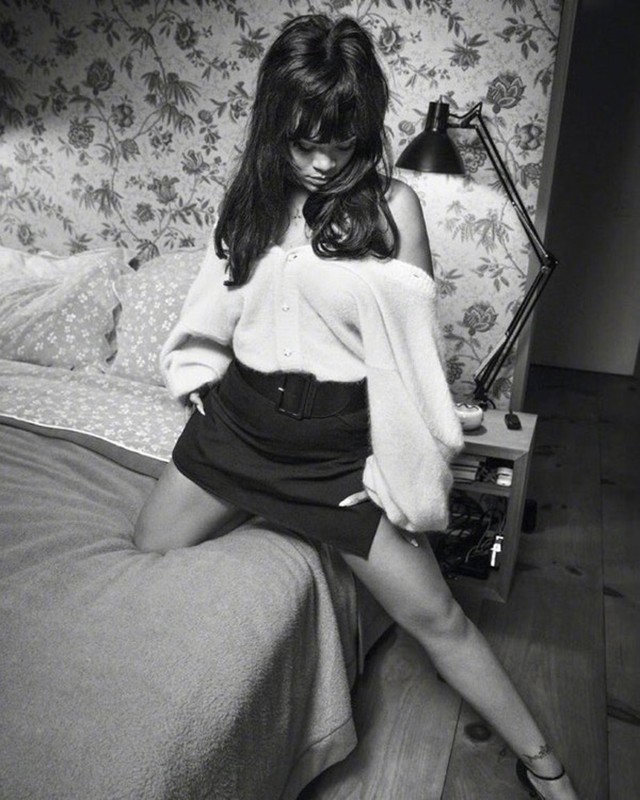 Portrait of a young woman with dark hair, wearing a cardigan and skirt, posing on a bed in front of a floral wallpaper.
