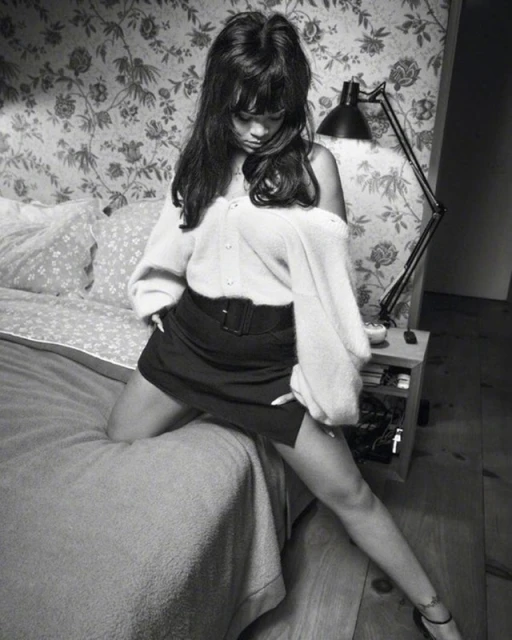 Portrait of a young woman with dark hair, wearing a cardigan and skirt, posing on a bed in front of a floral wallpaper.