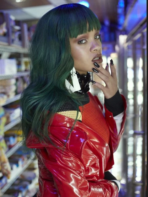 A woman with green hair, red coat, and black nails poses in front of shelves with grocery items in an indoor setting.