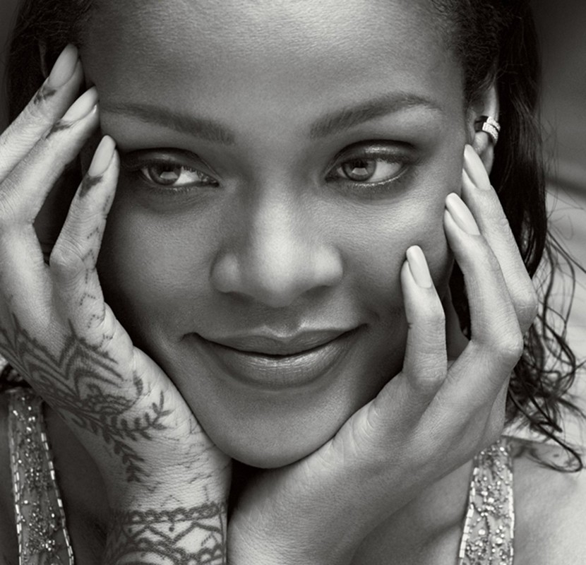 Close-up black and white portrait of Rihanna, smiling slightly with hands framing her face, showcasing henna tattoos on her hands.