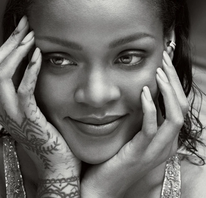 Close-up black and white portrait of Rihanna, smiling slightly with hands framing her face, showcasing henna tattoos on her hands.