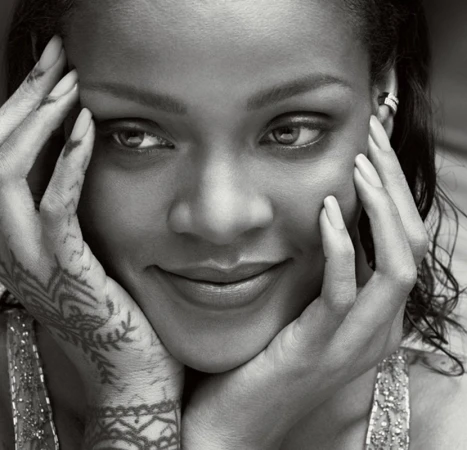 Close-up black and white portrait of Rihanna, smiling slightly with hands framing her face, showcasing henna tattoos on her hands.