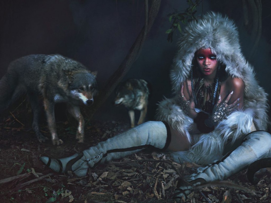 A woman in a fur hood and knee-high boots sits on a bed of leaves with two wolves in a dark, wooded outdoor environment.