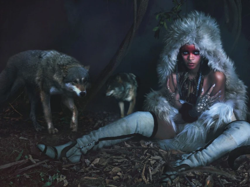 A woman in a fur hood and knee-high boots sits on a bed of leaves with two wolves in a dark, wooded outdoor environment.