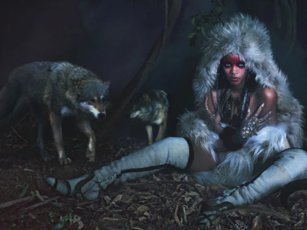 A woman in a fur hood and knee-high boots sits on a bed of leaves with two wolves in a dark, wooded outdoor environment.