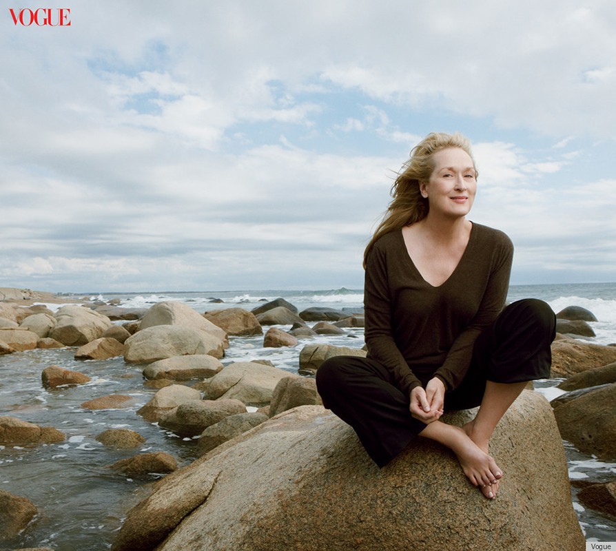 Meryl Streep poses on a rock at the beach for Vogue, wearing a long-sleeved brown shirt and dark pants, with ocean and sky in the background.