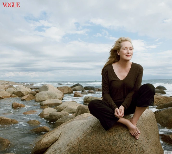 Meryl Streep poses on a rock at the beach for Vogue, wearing a long-sleeved brown shirt and dark pants, with ocean and sky in the background.