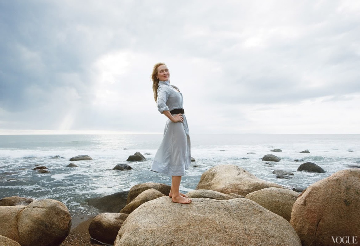 Blonde woman in a dress stands barefoot on a rock at the beach, gazing to the side with an overcast sky and ocean backdrop.