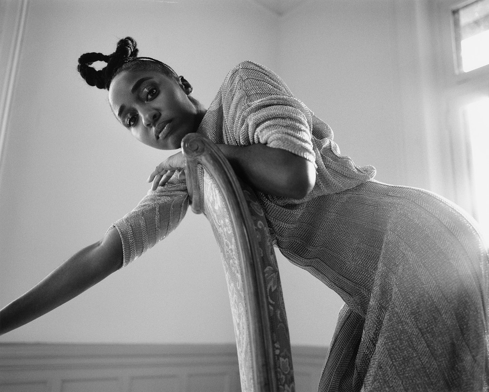 Eye-level portrait of African-American woman in shimmering dress leaning on antique chair in naturally lit room, against simple wall.