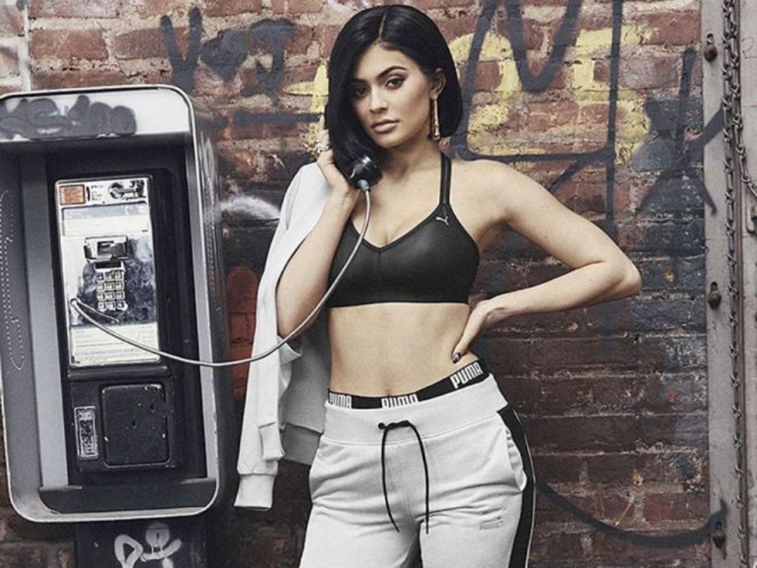 A woman in athletic wear poses with a payphone against a brick wall, casting a casual glance towards the camera.