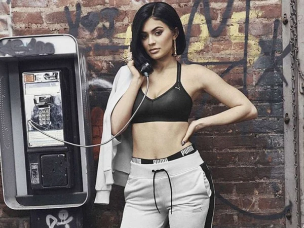 A woman in athletic wear poses with a payphone against a brick wall, casting a casual glance towards the camera.