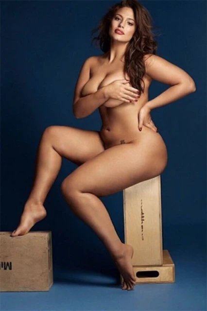 Plus-size model Ashley Graham poses nude in a studio setting, seated on a wooden box with a blue backdrop, showcasing body positivity.