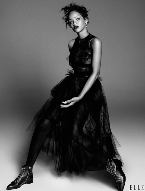 Monochrome portrait of Rihanna posing in a black tulle dress and combat boots against a grey backdrop in a studio setting. 