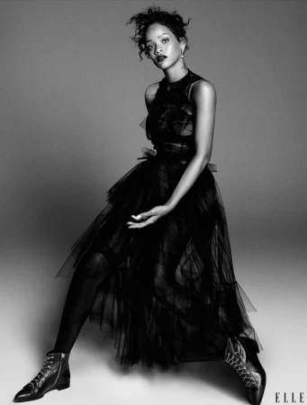 Monochrome portrait of Rihanna posing in a black tulle dress and combat boots against a grey backdrop in a studio setting. 