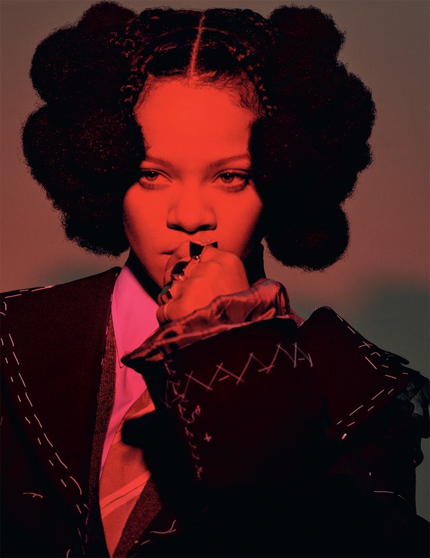 Portrait of a woman with large braided afro-style hair, wearing a black suit with white stitching, pink top and patterned tie.
