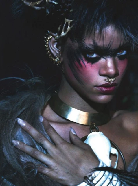 Close-up portrait of a model with creative makeup and jewelry, including a nose ring and collar necklace, set against a dark background.